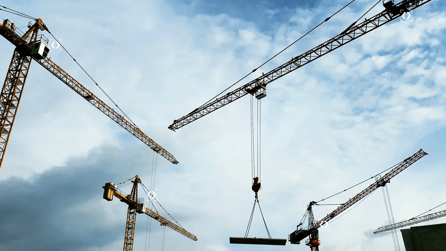 Tower cranes lifting a load against a cloudy sky, several framed by targeting markers